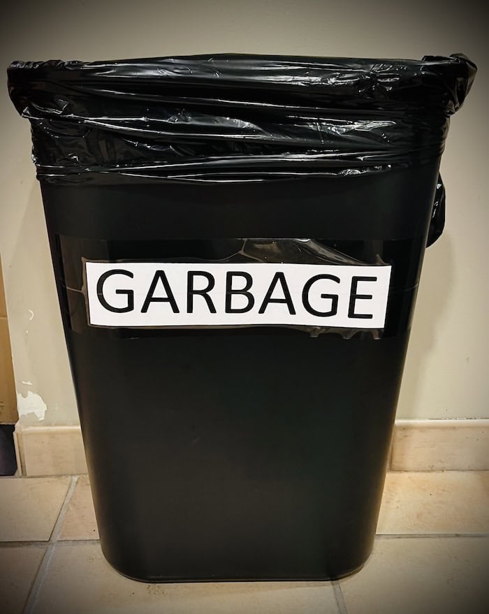 A black three gallon trash can with black liner bag labeled "Garbage," typed in all caps on white paper.